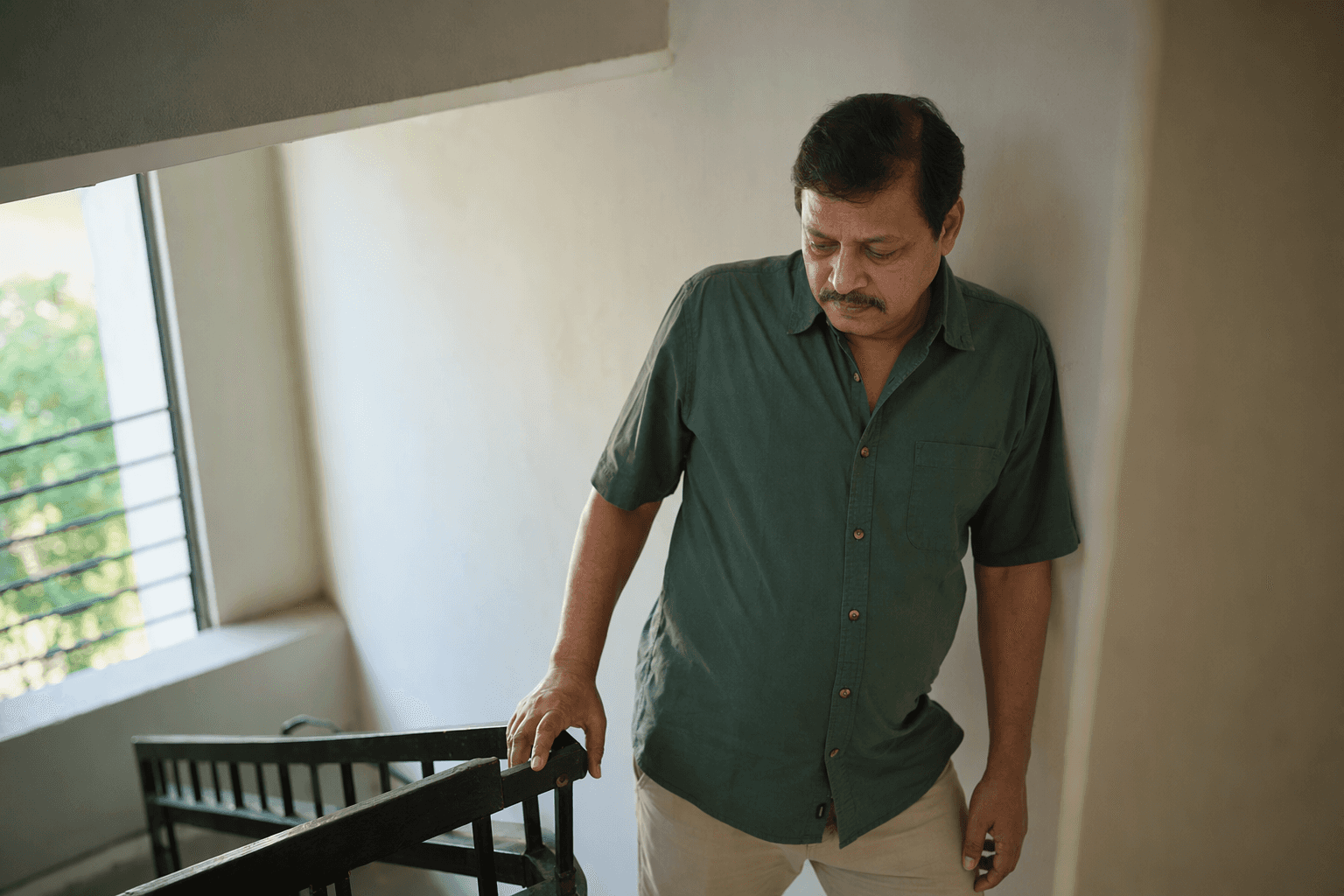 A man in his fifties pausing on a stairwell landing with a hand on the banister, catching his breath
