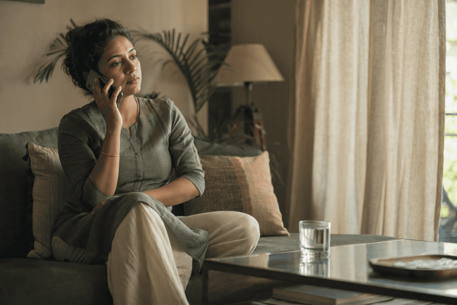 A woman in her thirties sitting on her sofa listening on the phone, late afternoon light through the curtain