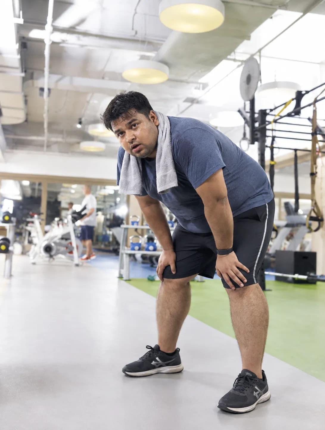 A man in a blue T-shirt and black shorts pausing in a gym with his hands on his knees and a towel round his neck, catching his breath between exercises