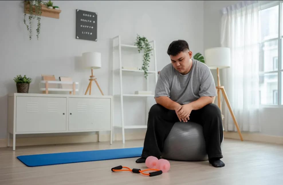A young man in a grey T-shirt sitting on an exercise ball in a bright living room, looking down at his hands, with a yoga mat, a resistance band and two small dumbbells on the floor