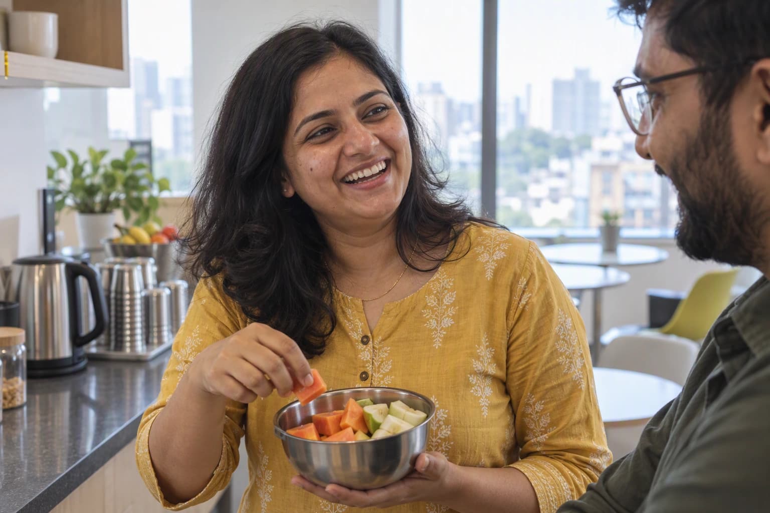 A woman laughs with a colleague in an office pantry, holding a bowl of cut fruit