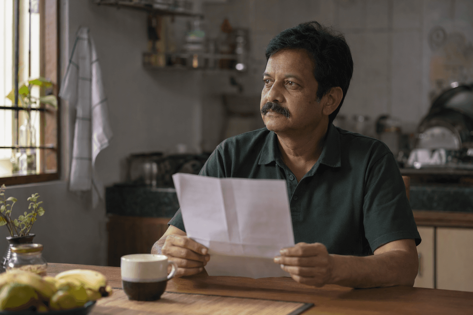 A man in his late forties at his kitchen table holding a folded printed report, looking away in thought