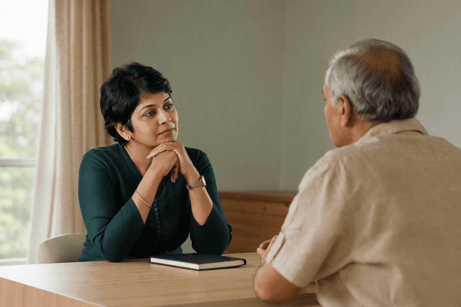 An Indian doctor in her forties listening to a patient across a small table in a bright, plain consulting room