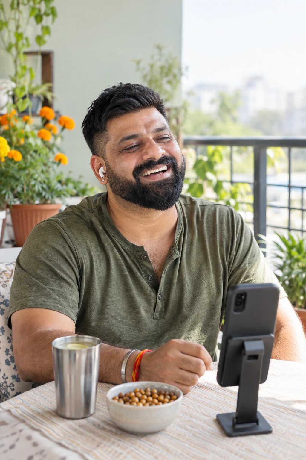 A bearded man in his early forties with a short textured crop laughing at a phone on a stand at his balcony table, earbuds in, an olive henley on