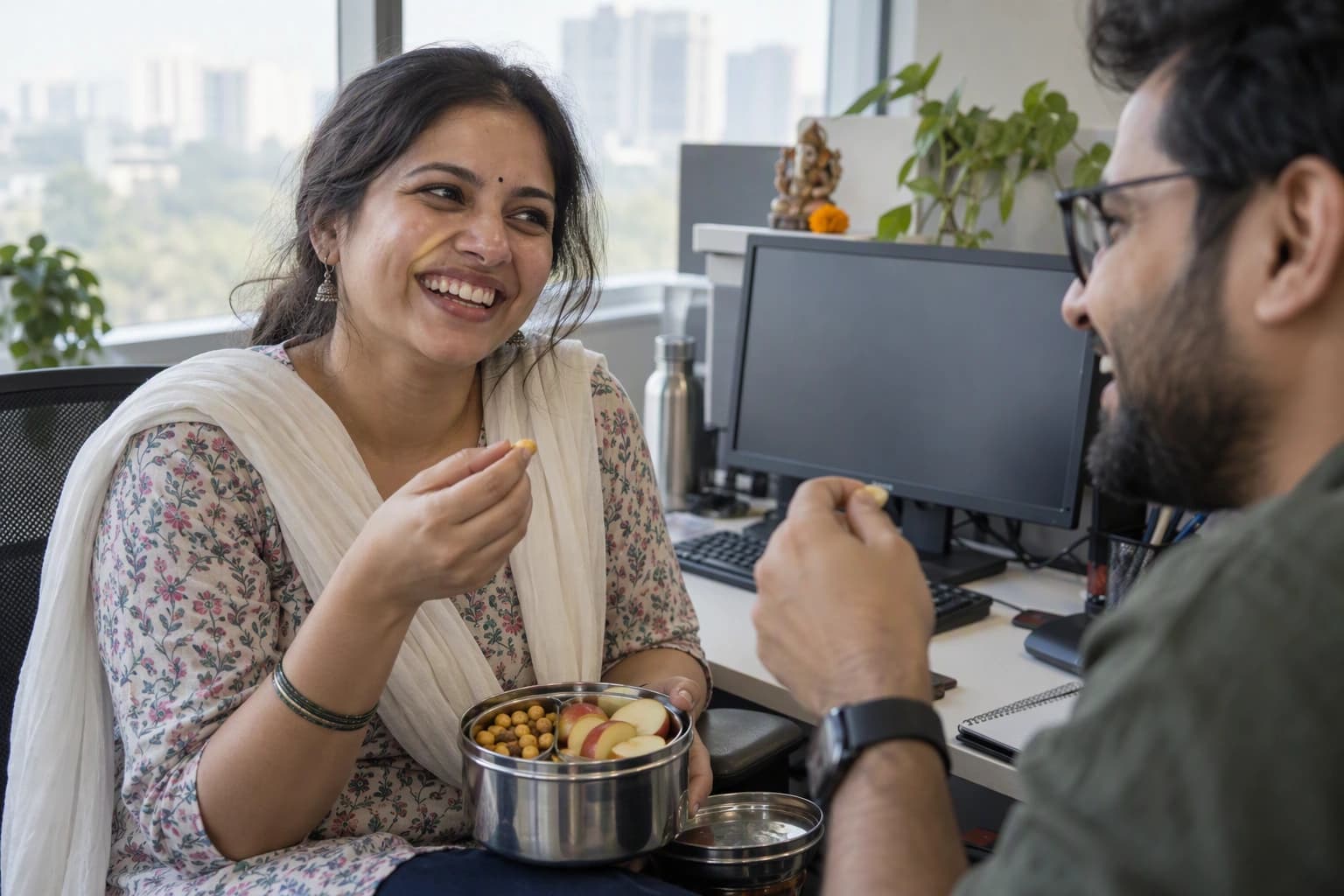 A woman laughs with a coworker as she shares roasted chana and apple slices at her desk
