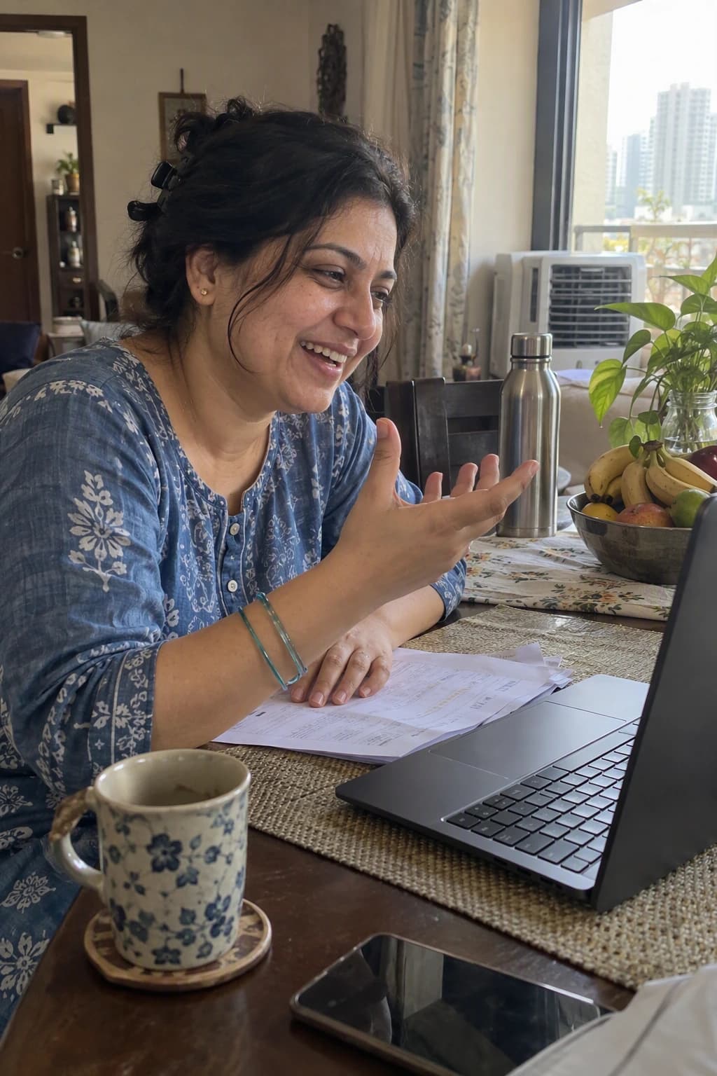 A woman at her dining table laughs during a video call on her laptop, a printed lab report under her hand