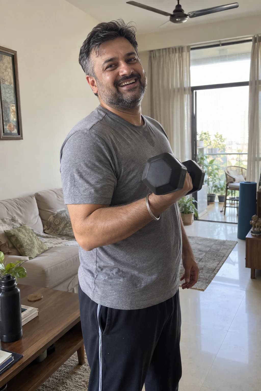 A man of about forty grins while doing a dumbbell curl in his Mumbai living room, a yoga mat and sofa behind him