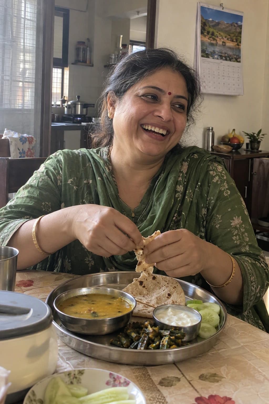 A woman in a green salwar kameez laughs as she tears a roti over a steel thali of dal, bhindi and curd at her home table